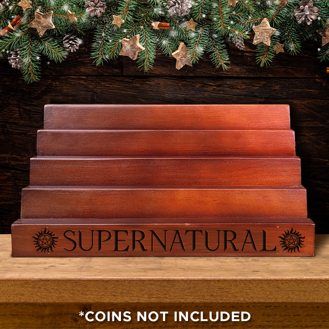 A wooden coin display rack with five tiers, on a table. Behind the rack is the bottom of a Christmas wreath. Black text on the bottom tier says "supernatural" with the anti-possession symbol at each side. 