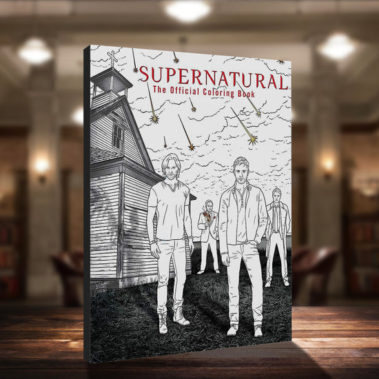 An image of a black and white book cover, standing on a wood table top in a library. At the front are Sam and Dean Winchester from the TV series Supernatural. Behind them are the characters Castiel and Crowley. They are all standing next to a church, and above them is a cloudy sky with star-shaped objects streaking toward the ground. At the top in red text is "Supernatural: The Official Coloring Book"