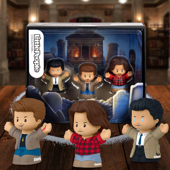 Three small Little People toy figurines on a wooden table, with the Bunker in the background. The figurines depict the characters Sam Winchester, Dean Winchester, and Castiel, from the TV series "Supernatural." Behind the figurines is a box containing three Little People toy figurines." The interior of the box depicts a cemetery, with an open grave at the front. A circle of salt surrounds the box.