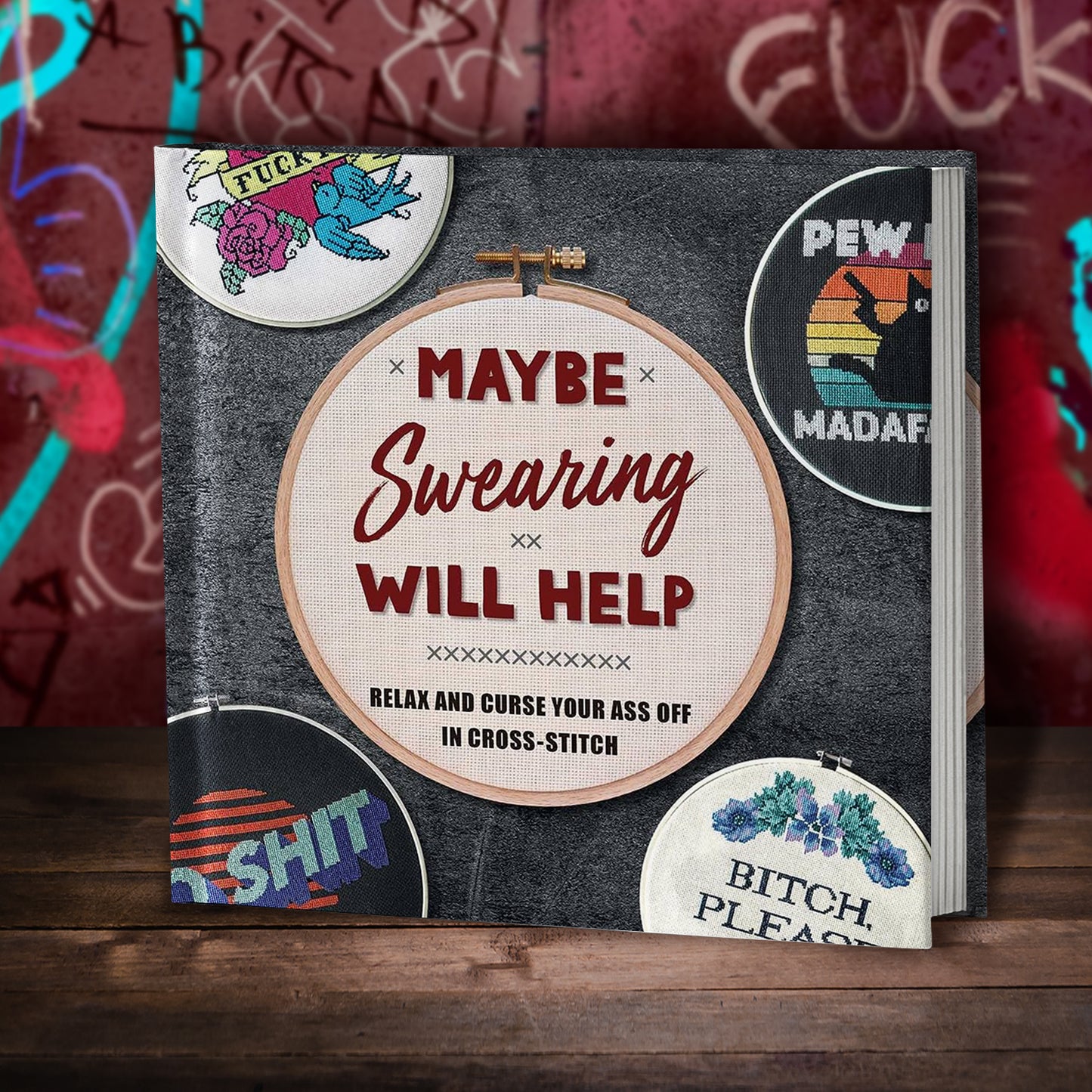 A black book on a wood table, with a graffiti-covered red wall behind it. On the cover is a round cross-stitch project with red text reading "Maybe swearing will help." Below that in black text is "relax and curse your ass off in cross-stitch."