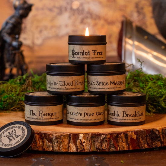 Six small candle tins, stacked in a pyramid on top of a cut piece of tree trunk. The labels all have names taken from the Lord of the Rings world. Behind the candles is a statue of a warrier, a piece of armor, and a blurred map of Middle Earth.