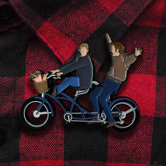 An enamel pin depicting Sam and Dean Winchester on a tandem bike. The pin is affixed to a red and black plaid shirt
