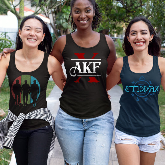 Three female models wearing tank tops, side by side. The left model's tank has the SPN 20 Years logo on the front. The center model's tank as the red and white AKF X logo. The model on the right has the Funnier in Enochian design. Behind them are trees along a path.