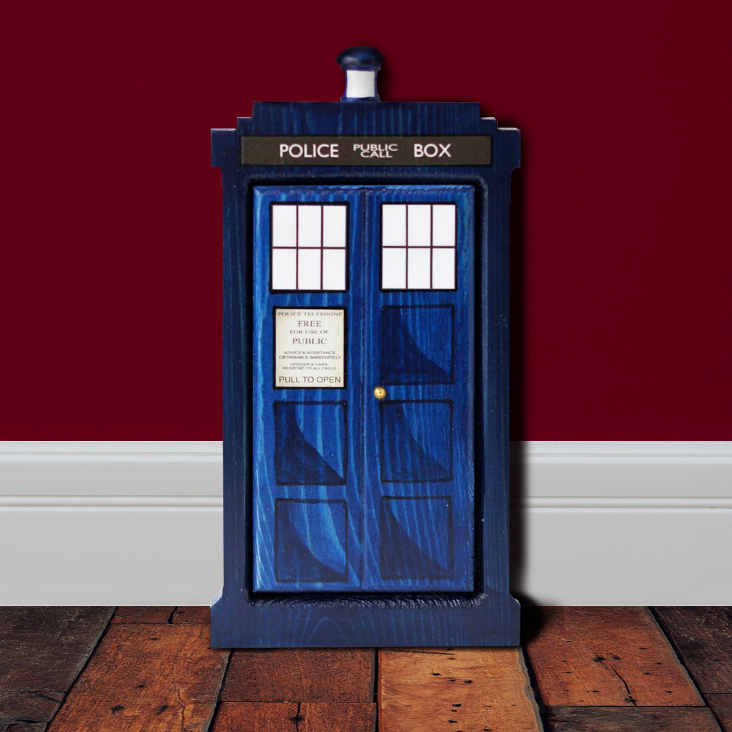 A front-facing rectangular wooden doorframe with a blue door, against a red wall and white baseboard. The door features the design of the TARDIS, including the POLICE BOX design, windows, and top light.