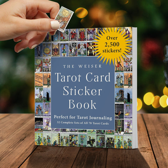 A photo of The Weiser Tarot Card Sticker Book, featuring rows of previews of Tarot card stickers in a traditional illustration design. The cover touts "Perfect for Tarot Journaling - 32 complete sets of all 78 Tarot cards - Over 2,500 stickers!". It is set against a green, holiday-light background and sitting on a wooden table. A woman's hand is seen holding a small sample sticker in front of it.
