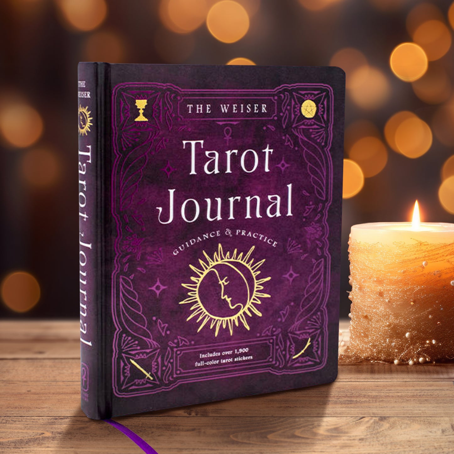 A purple book on a wooden table. The cover has purple sigils drawn across the front. White text in the center says “The Weiser Tarot Journal: guidance & practice.” Below the text is a yellow depiction of the sun, with a half-moon face in the center. Next to the book is a lit candle. In the background are twinkling amber-colored lights.