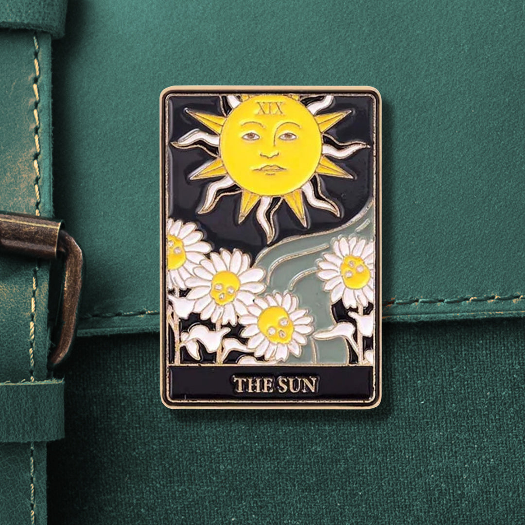 A rectangular tarot pin that reads "XIX" on top and "THE SUN" on the bottom. The pin depicts a large yellow-and-white sun with a face on the top. Beneath the sun is a grey river and 4 white daisies with yellow skulls in the center. 