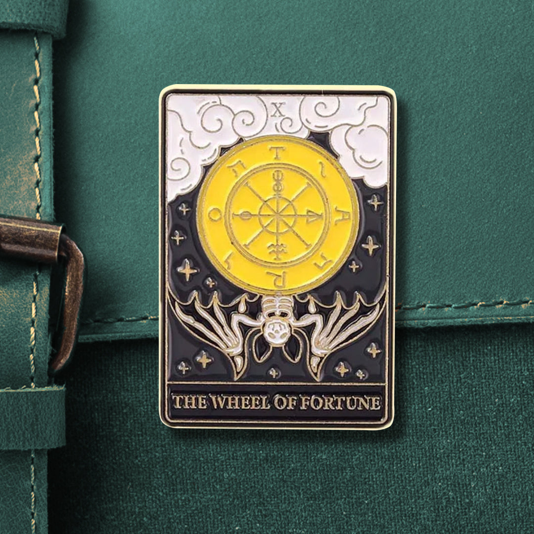 A rectangular tarot pin that reads the numeral "X" on top and "THE WHEEL OF FORTUNE" on the bottom, in gold. The pin depicts a yellow 'wheel of fortune' with a black-and-white skeleton bat hanging upside-down from the bottom of it. There are white, swirling clouds on the top and small gold stars around the background.