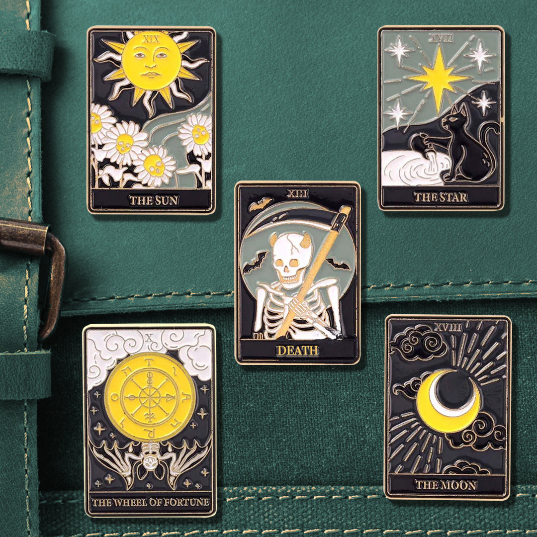 A close-up image of a set of five black enamel pins on an emerald green satchel. Each pin depicts a Major Arcana tarot card in black, grey, white, and yellow. The cards are Death, The Sun, The Moon, The Star, and the Wheel of Fortune.