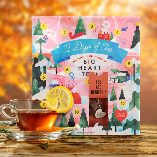 A multi-colored box against bright orange leaves. Various winter-themed drawings are across the front. At the top is a blue ribbon, with white text saying 12 days of tea. A glass teacup and saucer is next to the calendar, garnished with a slice of lemon.