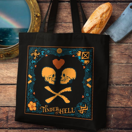 A black tote bag on a wooden table. The tote depicts two orange skulls facing each other, with a red heart above the,. Orange text at the bottom says "tender as hell." Around the border is a blue and orange pattern depicting horses, flowers, and fruits. Next to the bag is a portal, a loaf of bread, and a butcher's knife.