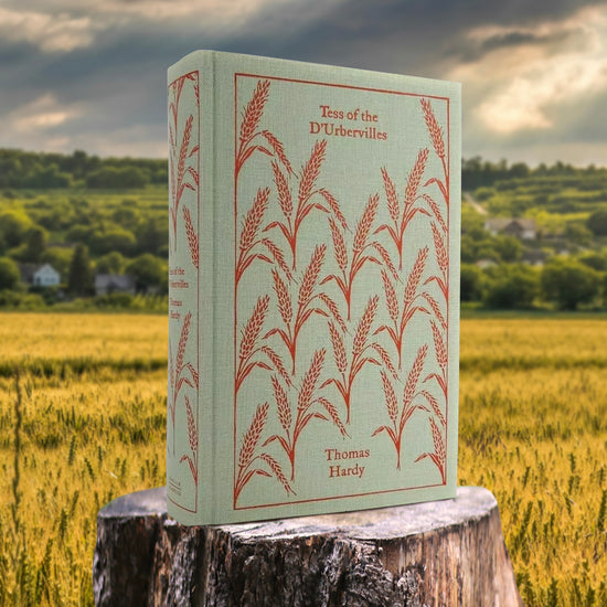 A sage green hardcover book on a tree stump, in the middle of a wheat field. The cover depicts drawings of wheat stalks in red. Red text says "Tess of the D'Urbervilles"