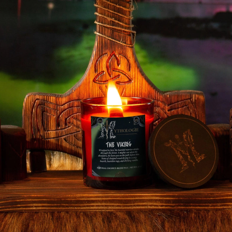 A glass candle on a wood table, with a green label. White text on the label says "The viking," with a desciption underneath. Next to the candle is a green lid. Behind the candle is a wooden Viking axe, against a swirling green and purple sky.