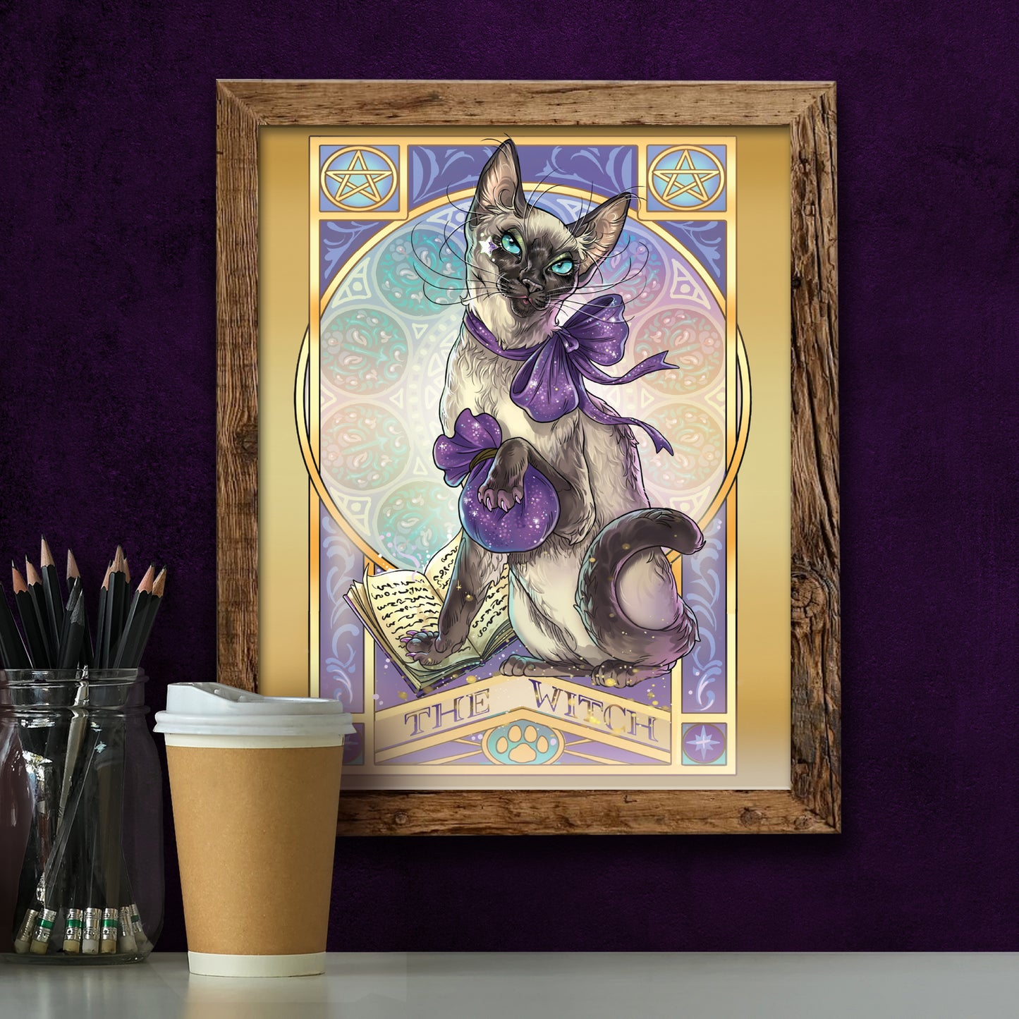 A framed drawing of a Siamese cat wearing a purple ribbon, and clutching a purple drawstring bag. The cat's front paw is on a spell book. In the background is a pattern of stained-glass style circles. At the top corners are yellow pentagrams. Purple text at the bottom says "The Witch." Below the drawing is a coffee cup and a glass jar filled with pencils.