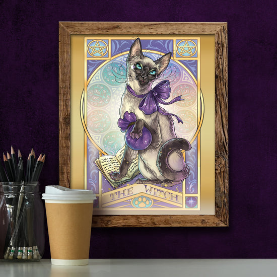 A framed drawing of a Siamese cat wearing a purple ribbon, and clutching a purple drawstring bag. The cat's front paw is on a spell book. In the background is a pattern of stained-glass style circles. At the top corners are yellow pentagrams. Purple text at the bottom says "The Witch." Below the drawing is a coffee cup and a glass jar filled with pencils.