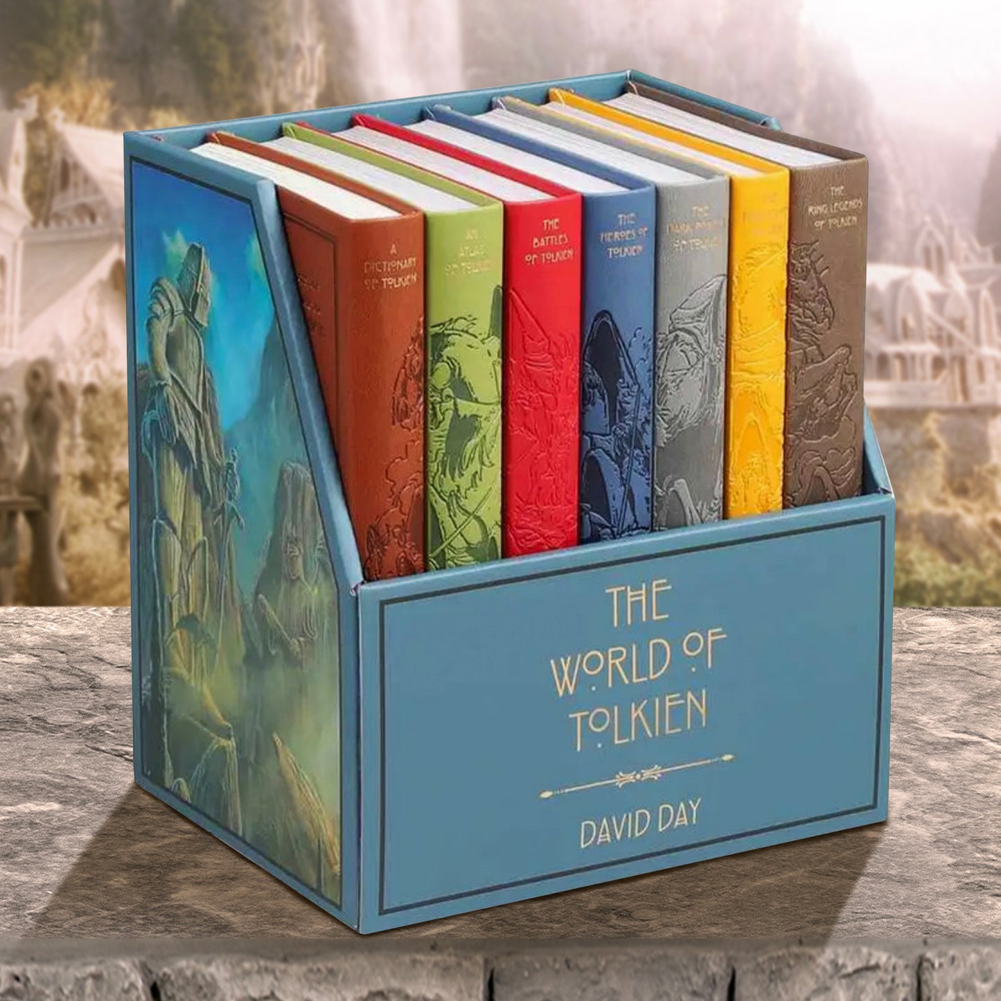 Seven hardcover books lined up side by side with their spines visible. Each book is a different color, with a drawing of creatures from the Lord of the Rings series spanning all seven books. The books are in a blue container with "The world of Tolkien" on the front in gold text. On the side of the box is a drawing of giant statues carved into cliffs. The book set is on a stone ledge, with an elven city in the background.