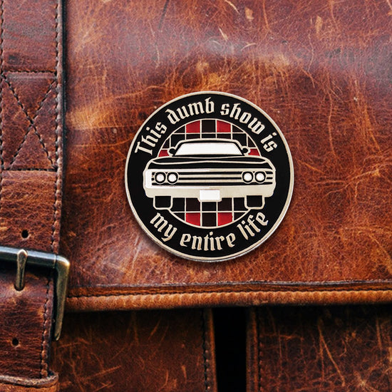 A circular black enamel pin that reads "THIS DUMB SHOW IS MY ENTIRE LIFE" around the edge. The center of the circle has a red-and-black checkered pattern and a vintage black Impala as seen from the front. The pin is fastened to a brown leather bag.