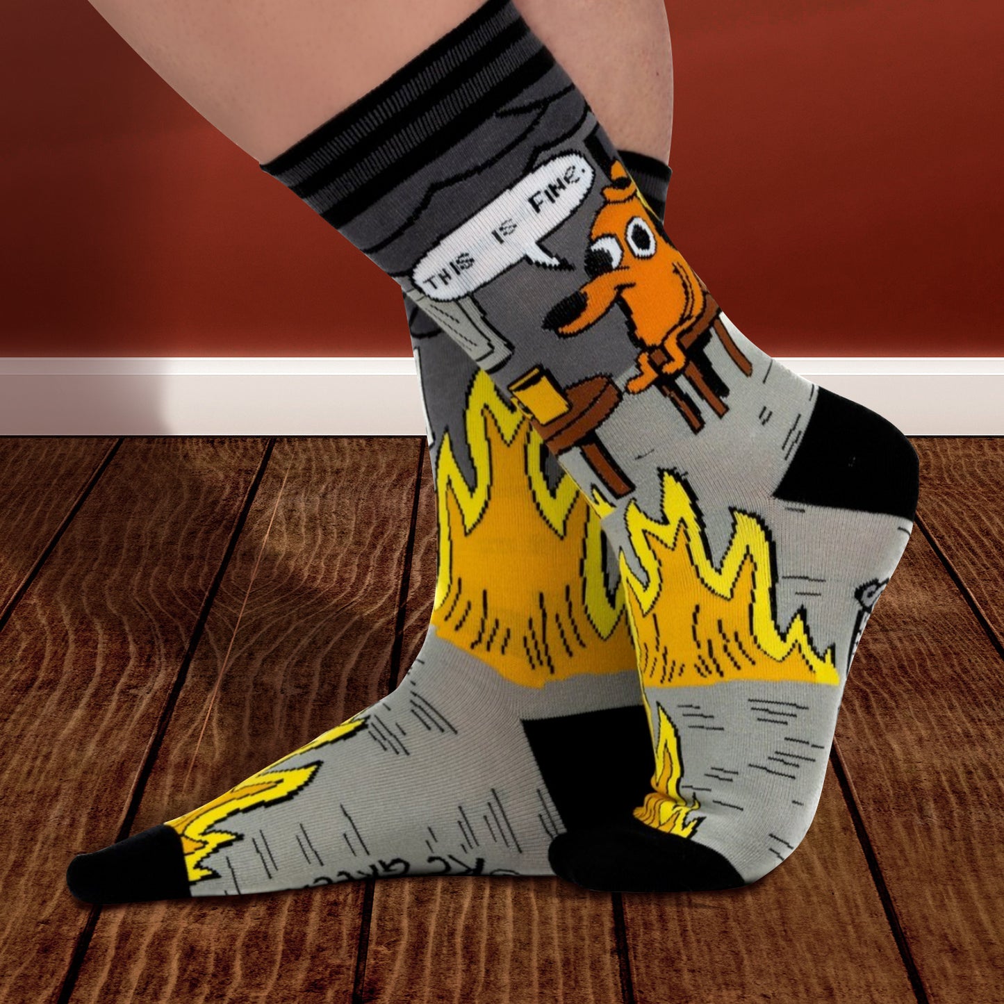 A model's feet wearing a pair of grey and back socks, on a wooden floor. One sock has a cartoon of a smiling brown dog wearing a hat, sitting in a chair in front of burning flames. Above him is a caption saying "This is fine." The other sock has a brown wooden table with a coffee cup, and a roaring flame underneath it. The cuff and toes of the socks are black. Behind the feet is a brown wall.