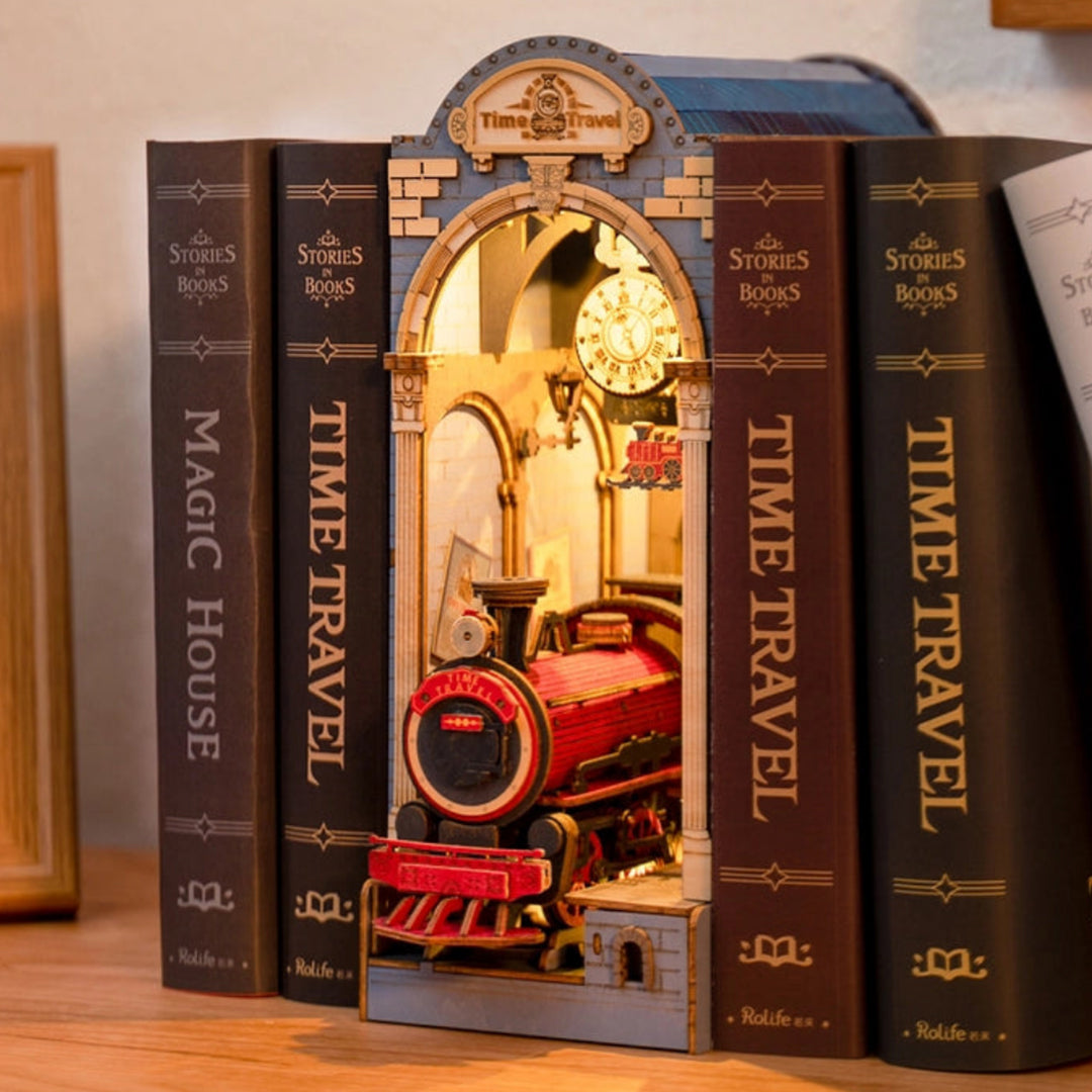 A 3/4-angle view of miniature train station with an open arched center, made of paper, on a bookshelf. The outside of the statio is light blue, with gold accents. At the top is a gold banner with "time travel" in dark gold text. A gold clock hangs from the top of the open arch. At the bottom is a red and black train locomotive, with "time travel" in yellow text on its front. Next to the station are books standing vertically.