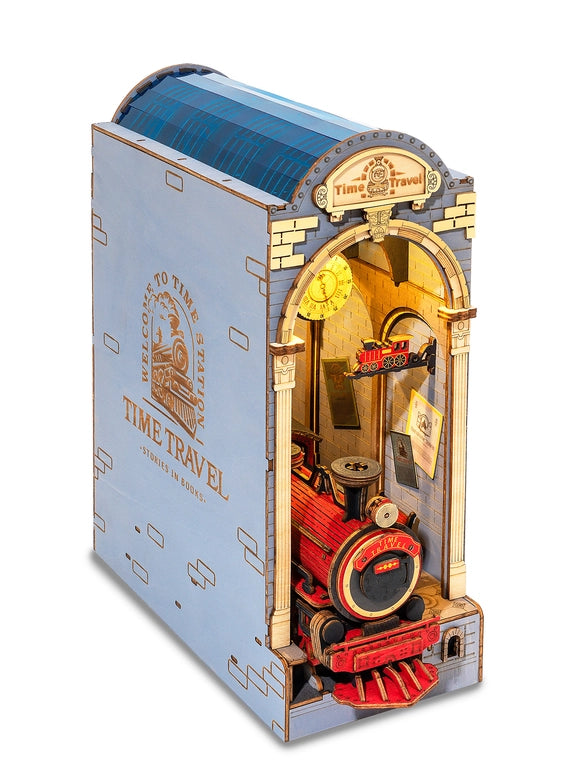 A miniature train station with an open arched center, made of paper. The outside of the statio is light blue, with gold accents. At the top is a gold banner with "time travel" in dark gold text. A gold clock hangs from the top of the open arch. At the bottom is a red and black train locomotive, with "time travel" in yellow text on its front. On the side of the station in gold is text saying "welcome to the station, time travel, stories in books," with a gold image of a train in the center.