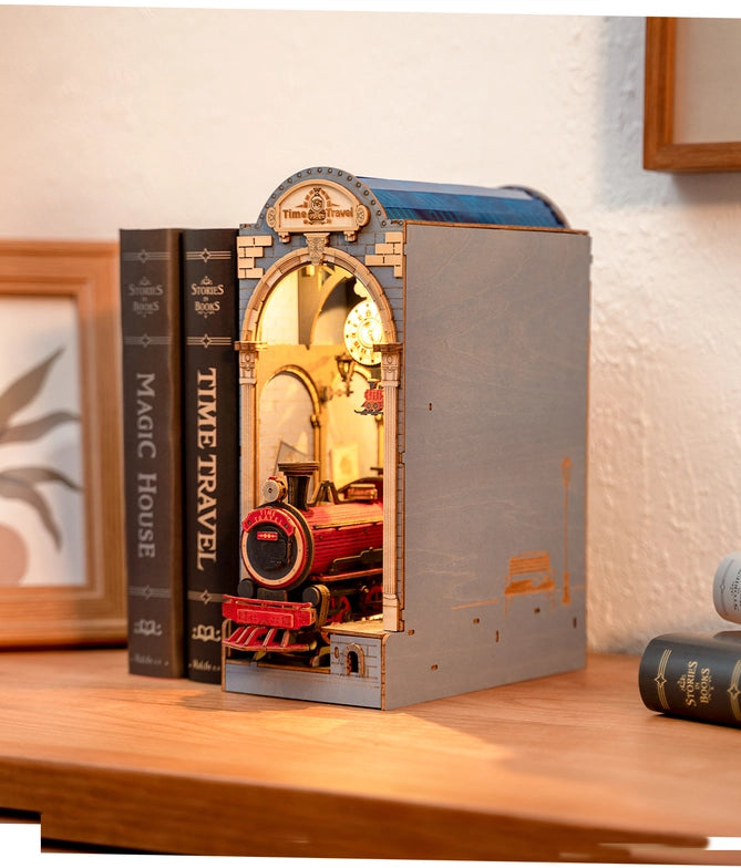 A miniature train station with an open arched center, made of paper, on a bookshelf. The outside of the statio is light blue, with gold accents. At the top is a gold banner with "time travel" in dark gold text. A gold clock hangs from the top of the open arch. At the bottom is a red and black train locomotive, with "time travel" in yellow text on its front. Next to the station are books standing vertically.