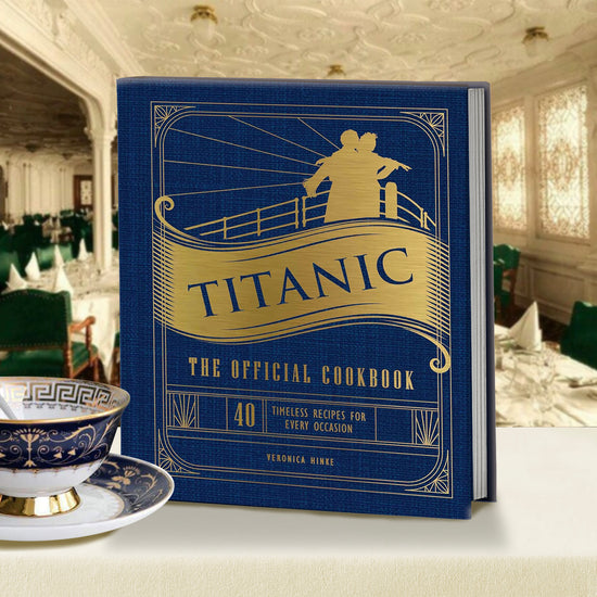 Load image into Gallery viewer, A blue book sitting on a fine dining table. The cover has gold text saying &amp;quot;titanic: the official cookbook.&amp;quot; A gold-colored silhouette of Jack and Rose from the movie &amp;quot;titanic&amp;quot; is at the top. There is a blue and white saucer and tea cup sitting to the left of the cookbook.

