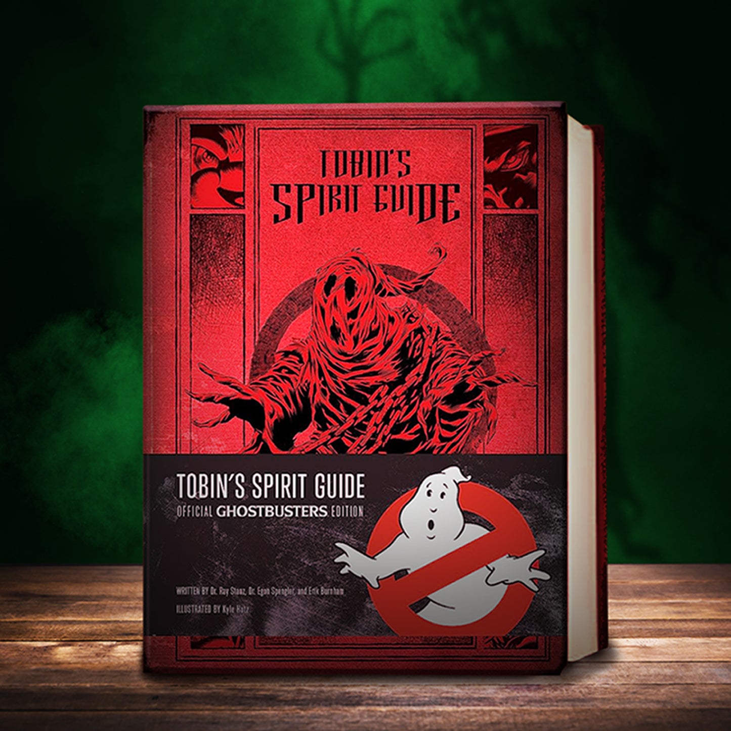 A red book cover with the title "Tobin's spirit guide" in black text. In the center is a black drawing of a creepy robed ghost. At the bottom of the cover is a black band with the book title, and the Ghostbusters logo on the right side. The book is sitting on a wood table top, in front of a smoky green and black background.