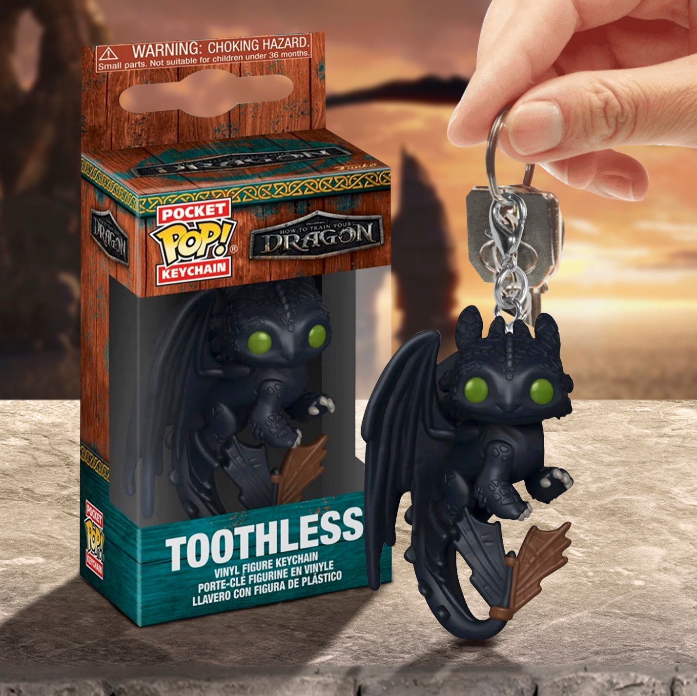 A model's hand holding black keychain depicting the dragon Toothless from the live action movie "How to Train Your Dragon." The dragon has dark green eyes and claws, and has a metal key ring on its head. Next to the keychain is a cardboard box depicting the character. In the background is an orange sunset. Under the items is a stone ledge.