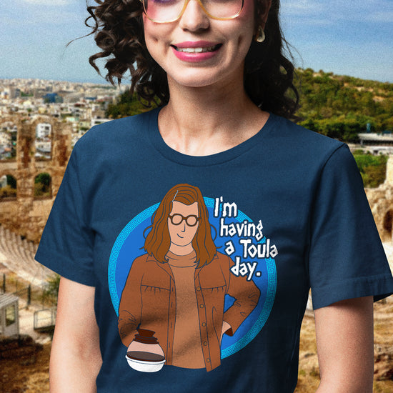 Person wearing a navy blue t-shirt with a graphic and text 'I'm having a Toula day.' in front of a scenic background.