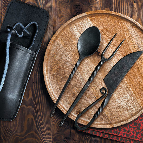 A spoon, form, and knife on a wooden plate, all made of black iron, on a wooden plate. Each has a decorative twist along the length of its shaft. The fork has two prongs, and the knife's handle folds back on itself. Next to the plate is a black leather holder for the cutlery. Below the plate is a sprig from a green tree, a red napkin.