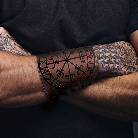 Close up of a male model's crossed forearms, against a black T-shirt. On the model's right forearm is a wide, brown leather wrist cuff, adorned with Nordic runes. The model's hands feature black tattoos of floral and geometric patterns.