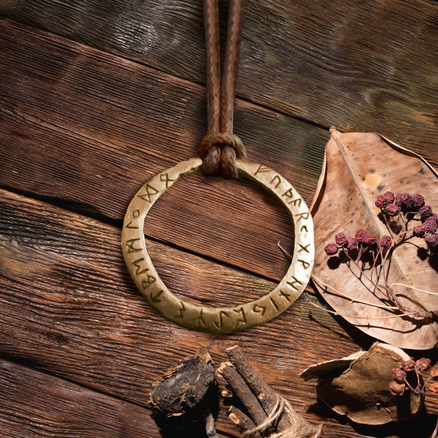 Close up of a brass pendant on a rope necklace, against a wooden tabletop. The pendant is an open circle, with Nordic runes printed around the edge. Next to the pendant are dried leaves and berries.