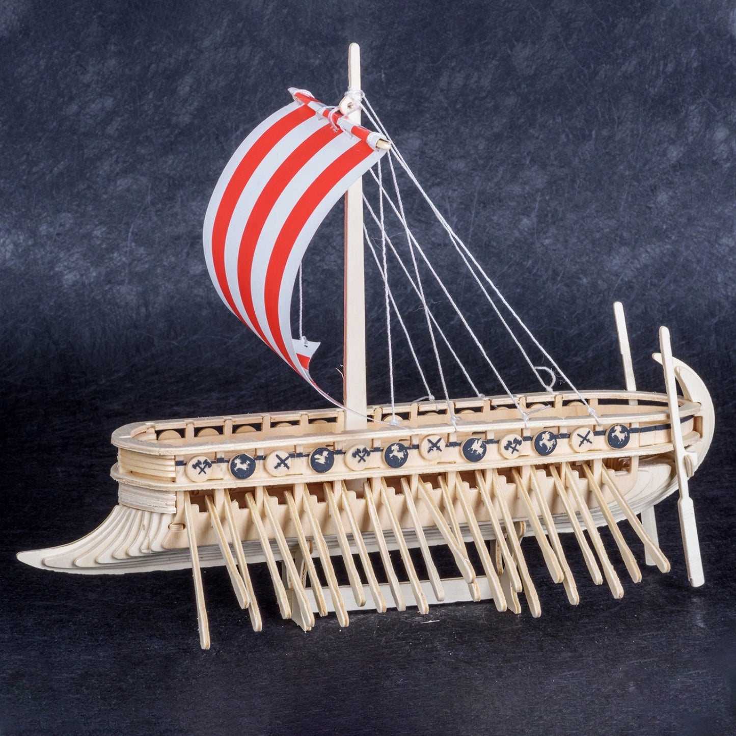Side view of a miniature wooden Viking ship with a read and white sail, set against a dark blue background. The ship has many oars sticking out the sides, and miniature wooden shields line the edge, with drawings of axes and dragon heads on each.