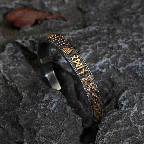 A black metal bracelet standing in a rock crevice. The bracelet features Nordic runes and symbols in gold.