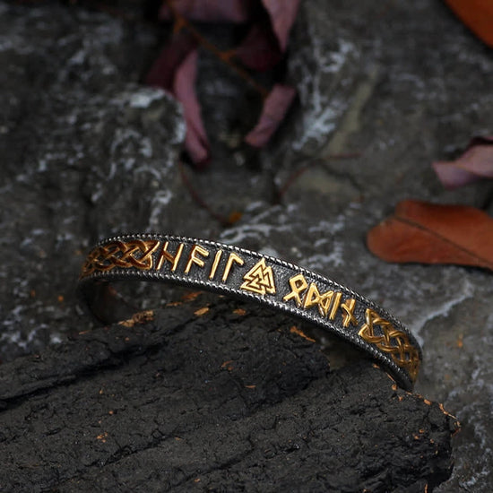 A black metal bracelet standing in a rock crevice. The bracelet features Nordic runes and symbols in gold.