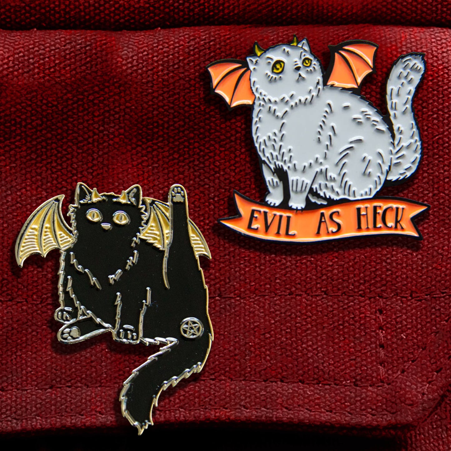 Two enamel pins attached to a red canvas bag. One pin is in the shape of a black cat, with yellow bat-like wings. One of the cat’s hind legs is lifted, and the cat’s butthole is covered by a pentagram. The other pin is in the shape of a grey cat with orange bat wings and yellow horns. Under the cat is an orange ribbon with black text saying "Evil as heck."