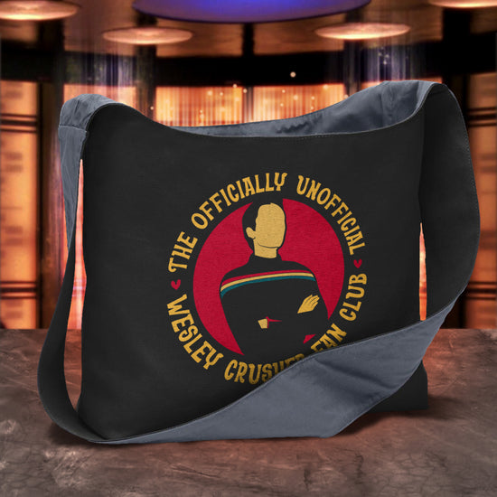 A black cross-body sling bag sitting on a stone table, with a Star Trek transporter room in the background. The front of the sling bag depicts a figure in a dark shirt with arms crossed, and a rainbow stripe across the chest. Yellow text around the image says "The unofficially official Wesley crusher fan club." 