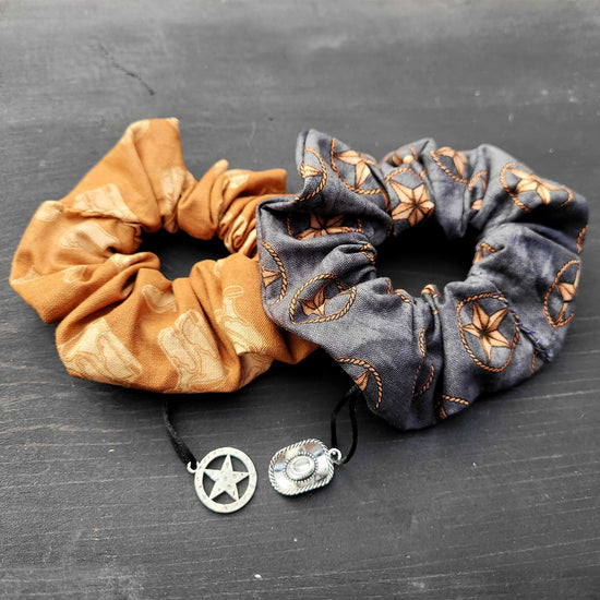 Two scrunchies lay side by side on a black wooden table. The scrunchie on the left is a burnt orange with a beige cowboy boot print and a Texas Ranger badge charm. The one on the right is a grey-black color with a print of brown ranger stars ringed with rope, and has a cowboy hat charm.