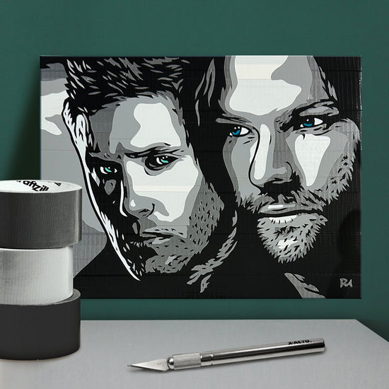 A black and white image of Sam and Dean Winchester, created with different shades of duct tape. The piece is resting against a pine green background, and 3 rolls of duct tape and an Xacto knife are visible on the table below.