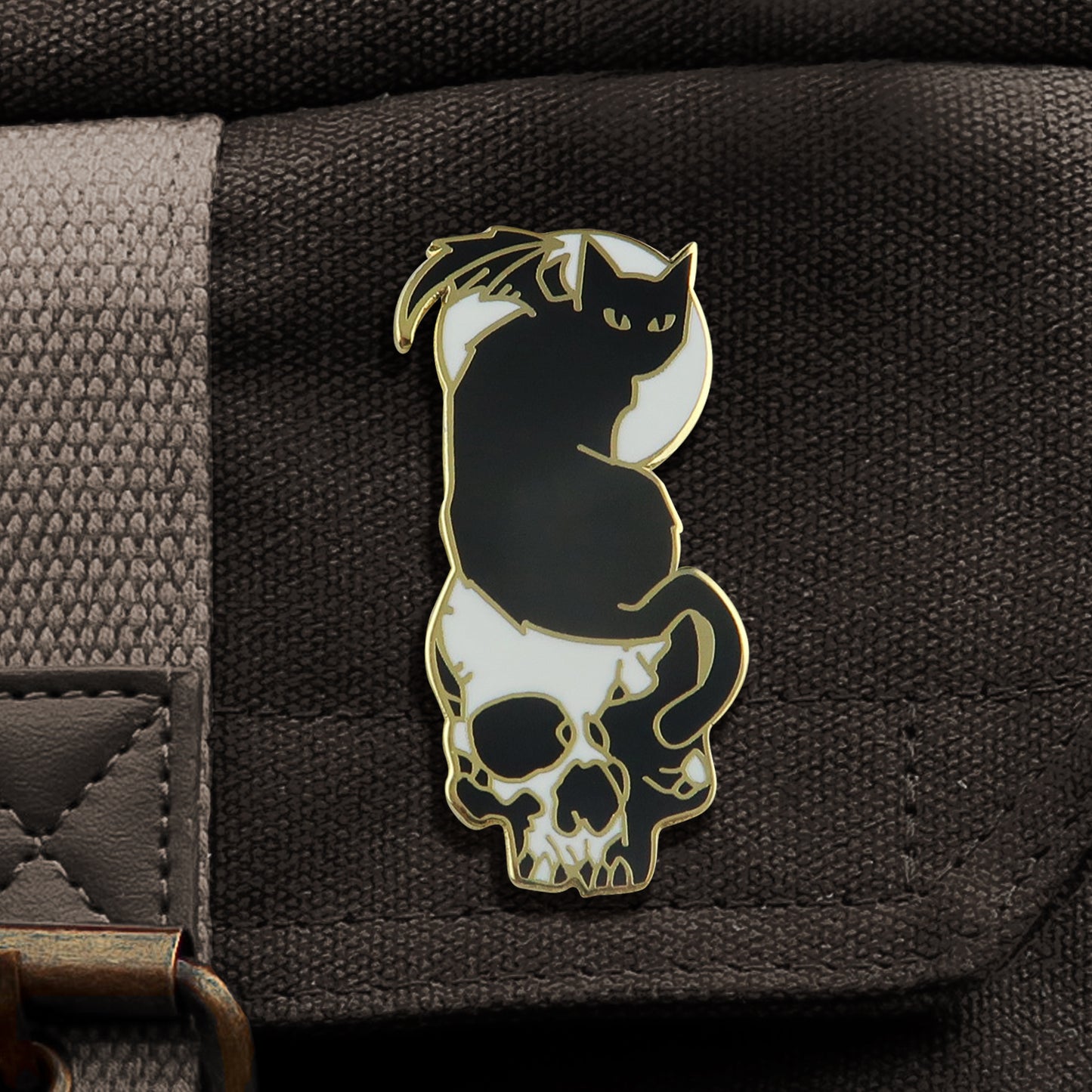 A black and gold enamal pin, attached to a black canvas bag. The pin depicts a black cat with bat wings, seen from the back with its head facing behind it. The cat is sitting on top of a white skull, and heind the cat is a round white moon. The cat's tail hangs down over the skull's left eye.