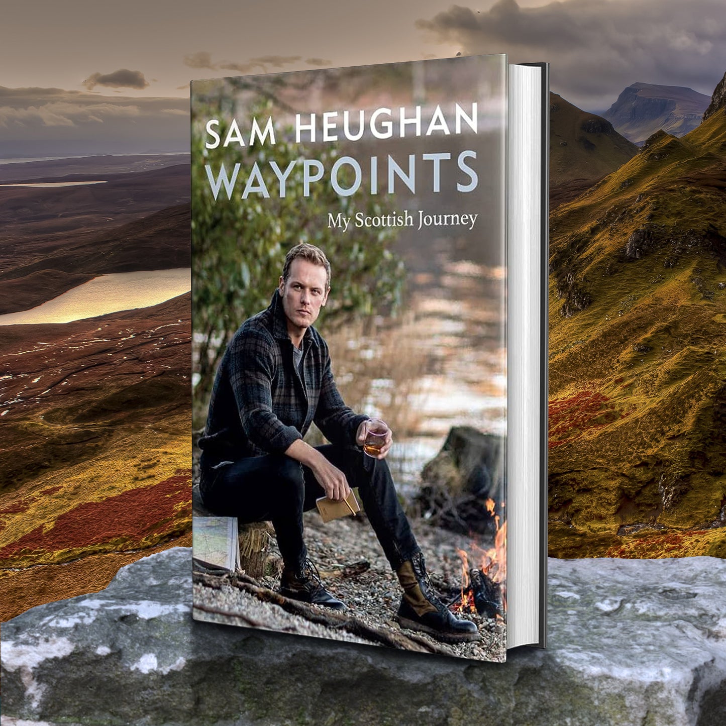 An image of a book standing on a flat rock, in front of a mountain scape. On the front of the book cover is actor Sam Heughan, sitting on a rock in front of a River. At the top in white text is "Sam Heughan: Waypoints, my scottish journey."