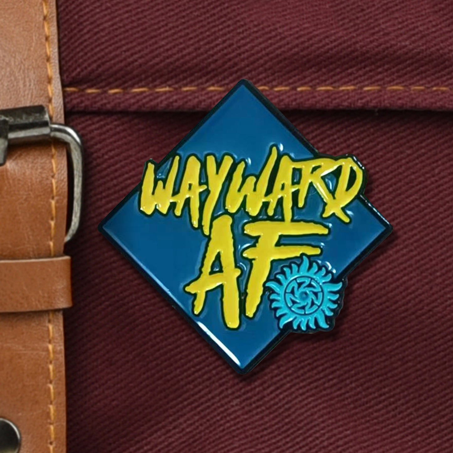 A diamond-shaped pin with a navy blue background and yellow text reading "Wayward AF" with a teal symbol in the corner. The pin is attached to a deep red canvas bag with a brown leather buckle strap.