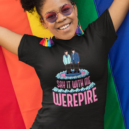 A female model wearing a black t-shirt, against a rainbow flag background. On the front of the shirt is pink text that says "say it with me, werepire." Above the text is a depiction of a wedding cake topper with two grooms holding hands.