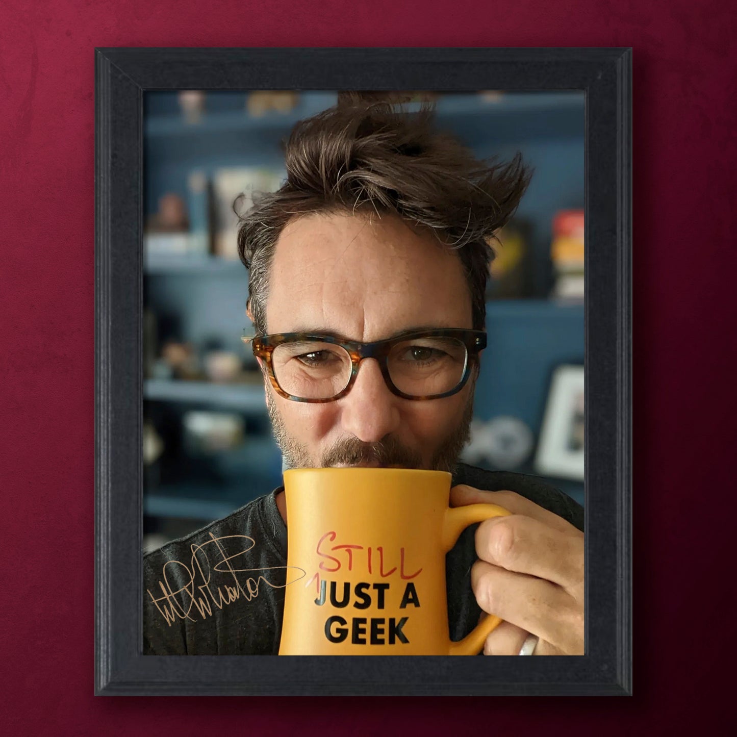 Framed picture of actor Wil Wheaton holding a mug with 'Still Just a Geek' text, against a blurred indoor background.