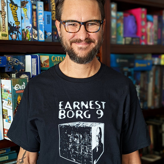 Actor Wil Wheaton standing in front of a shelf filled with board and card games. He's wearing a black crew neck t-shirt that reads EARNEST BORG 9 and has an illustration of the Borg Cube beneath in white print.