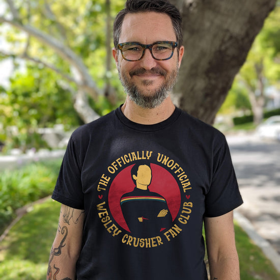 An image of actor Wil Wheaton in a black T-shirt. The front of the T-shirt depicts a figure in a dark shirt with arms crossed, and a rainbow stripe across the chest. Yellow text around the image says "The unofficially official Wesley crusher fan club."