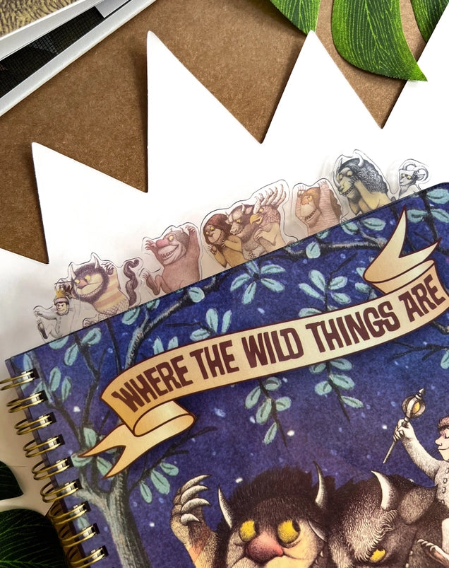 A blue journal depicting Max and two monsters from Where the Wild Things Are. Two monsters are carrying Max on their shoulders. At the top is a banner across two trees with text saying where the wild things are. At the top of the journal are tabs depicting characters from the book.
