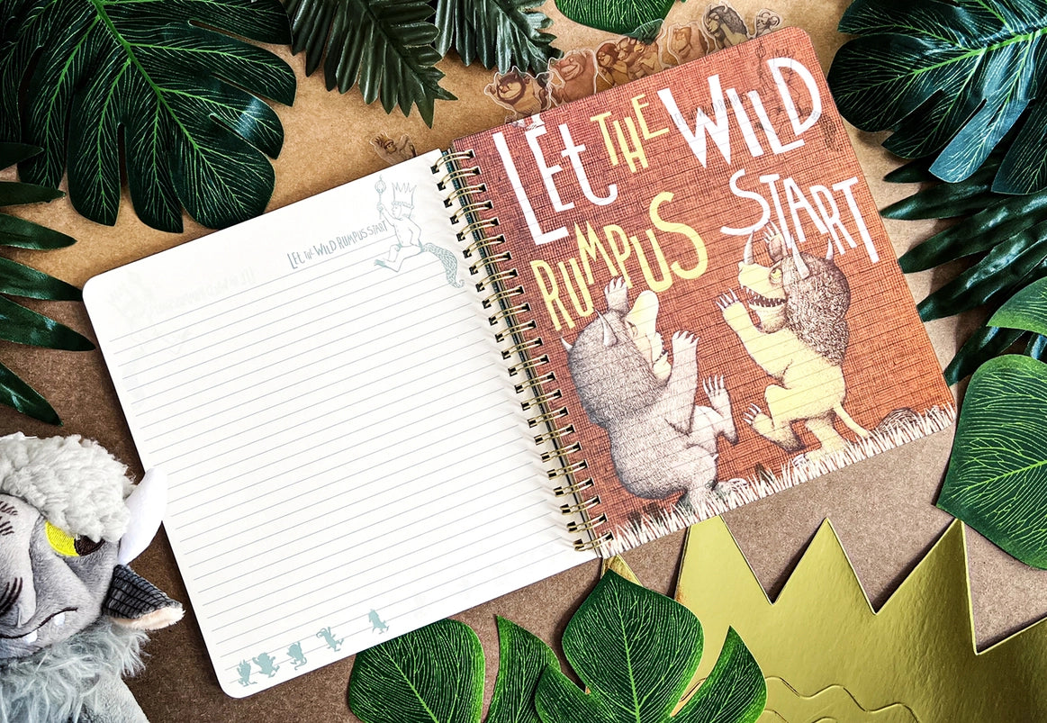 A page from the book showing two monsters against a red background. White and yellow text says "let the wild rumpus start." The journal is on a table surrounded by leaves and a goat puppet.