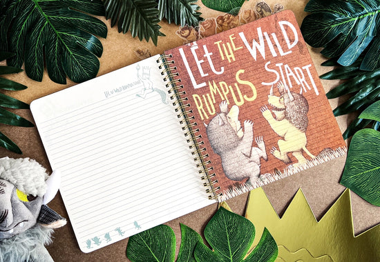 A page from the book showing two monsters against a red background. White and yellow text says "let the wild rumpus start." The journal is on a table surrounded by leaves and a goat puppet.