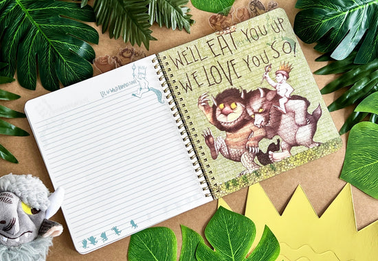 A page from the book showing two monsters carrying Max, against a yellow background. brown text says "we'll eat you up we love you so." The journal is on a table surrounded by leaves and a goat puppet.